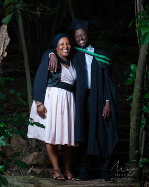 Two people in graduation gowns and attire share a joyful moment together in a wooded outdoor setting.