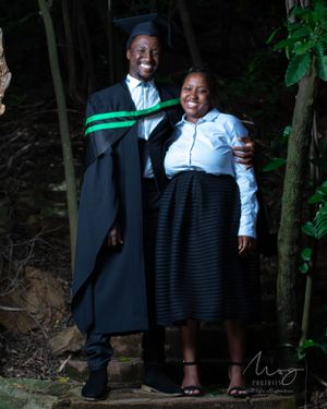 Two people stand together outdoors, one wearing a graduation gown with green sash while the other wears a white shirt and skirt.