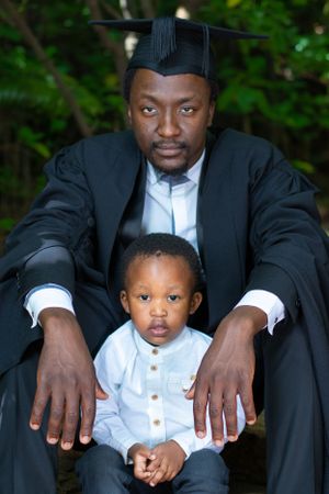 Graduate in cap and gown poses with small child wearing white shirt in outdoor setting.