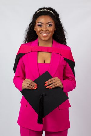 Graduate in bright pink suit holding graduation cap with tassel against light background.