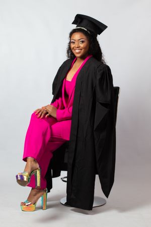 Graduate in bright pink jumpsuit and black academic regalia poses in studio with stylish platform heels.