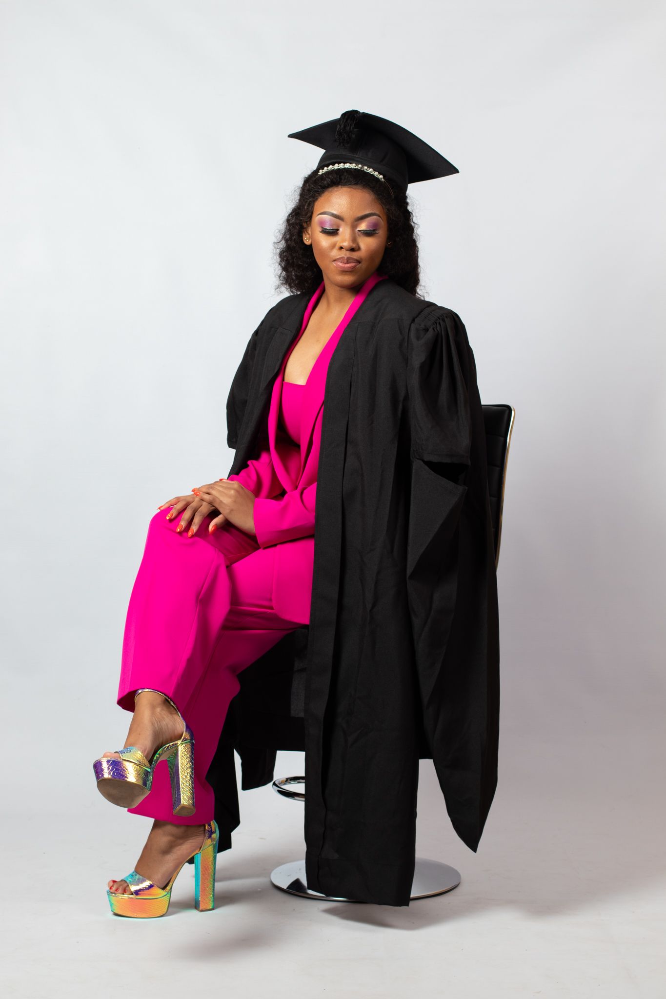 Graduate in black cap and gown with bright pink jumpsuit poses elegantly in chair against white backdrop.