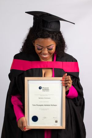 Graduate in cap and gown holds diploma while smiling against white background.