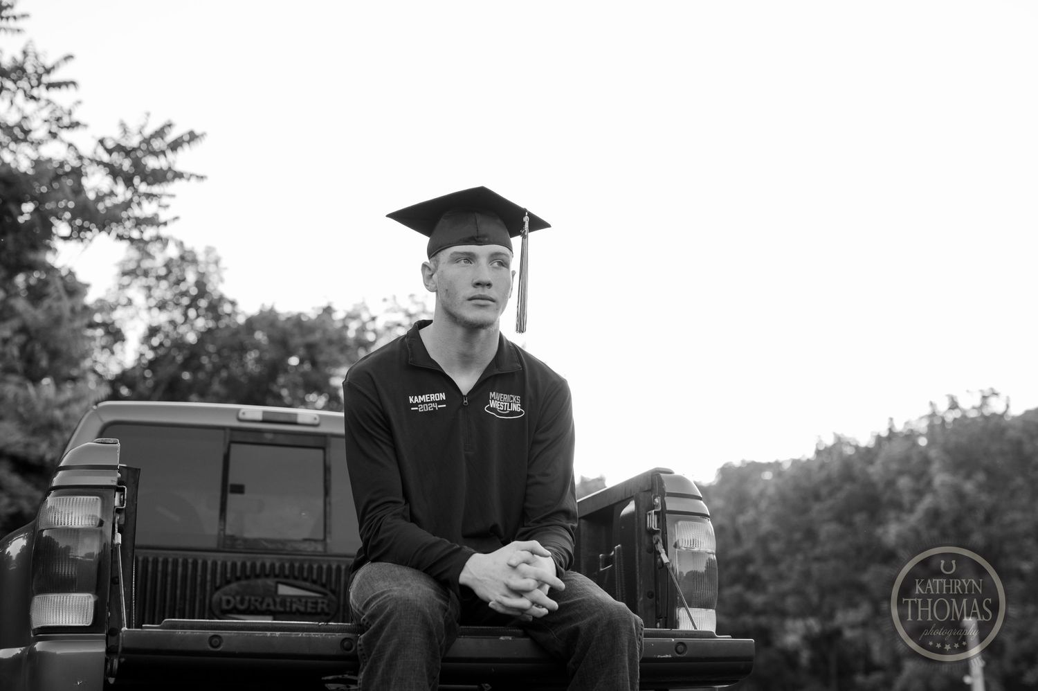 Black and white photo of graduate sitting on truck tailgate wearing cap and polo.