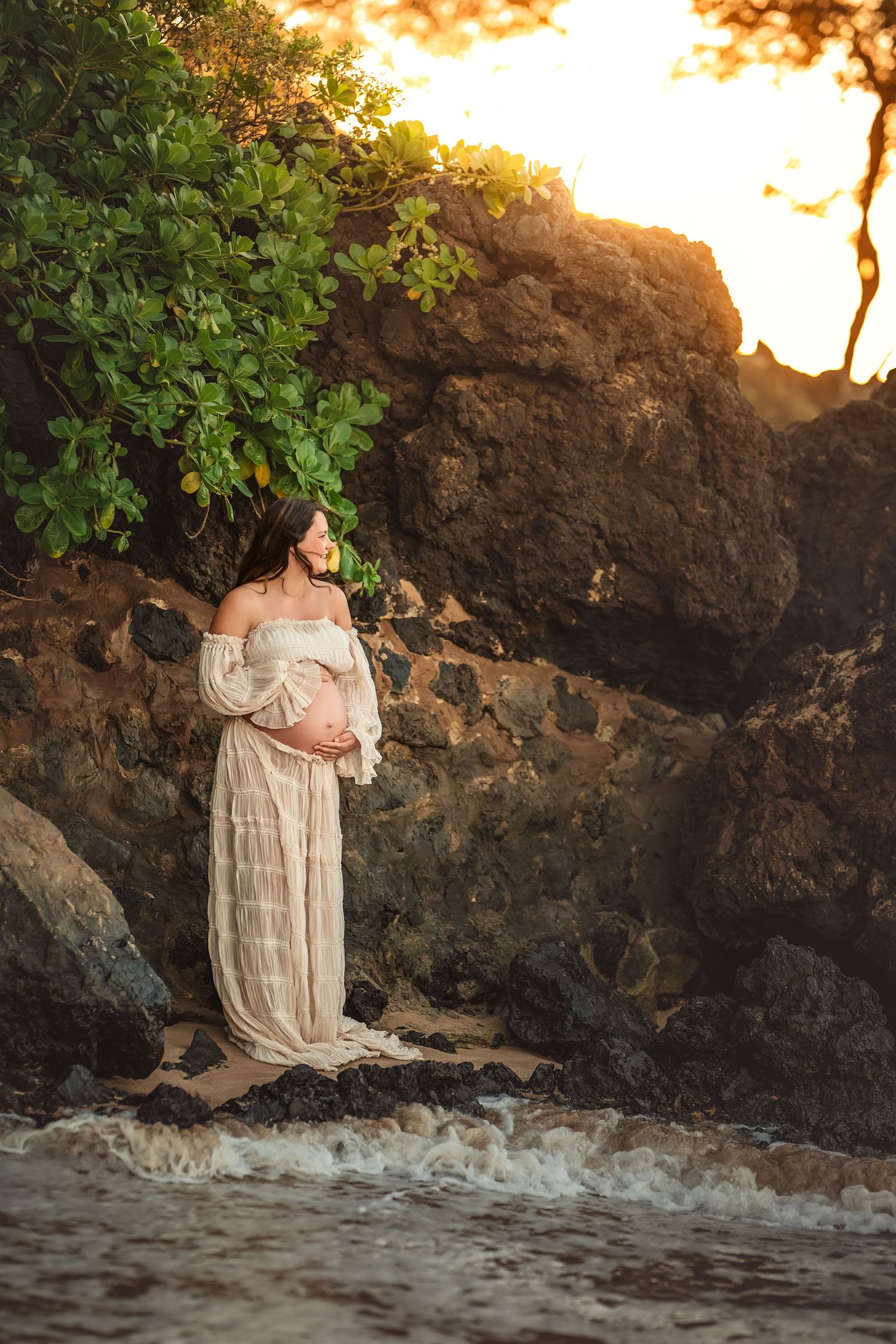 A pregnant woman in a flowing cream two-piece dress poses on a sandy beach at sunset against dramatic rocky cliffs and lush foliage.