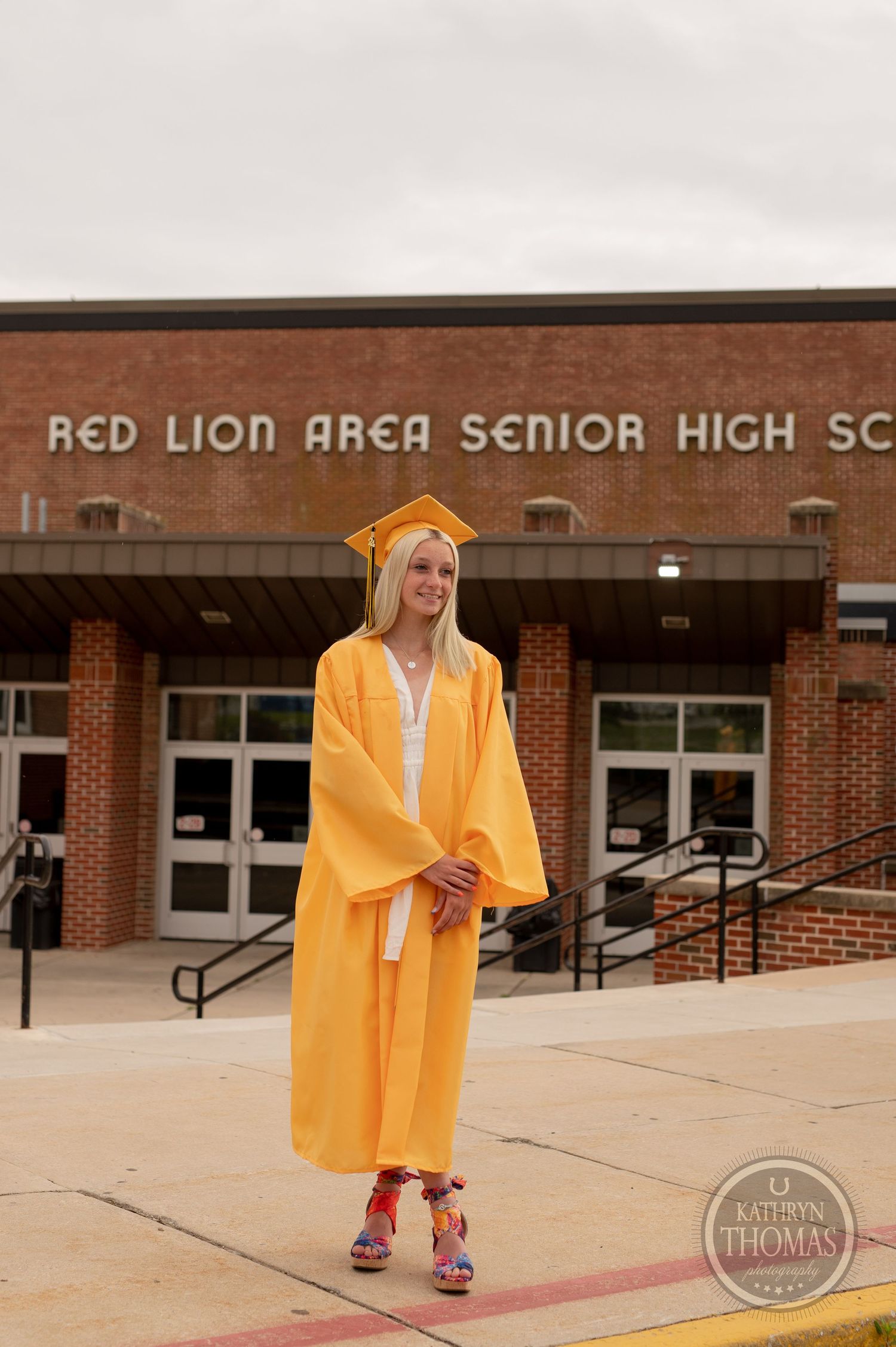 Graduate wearing yellow cap and gown stands outside brick school building entrance.