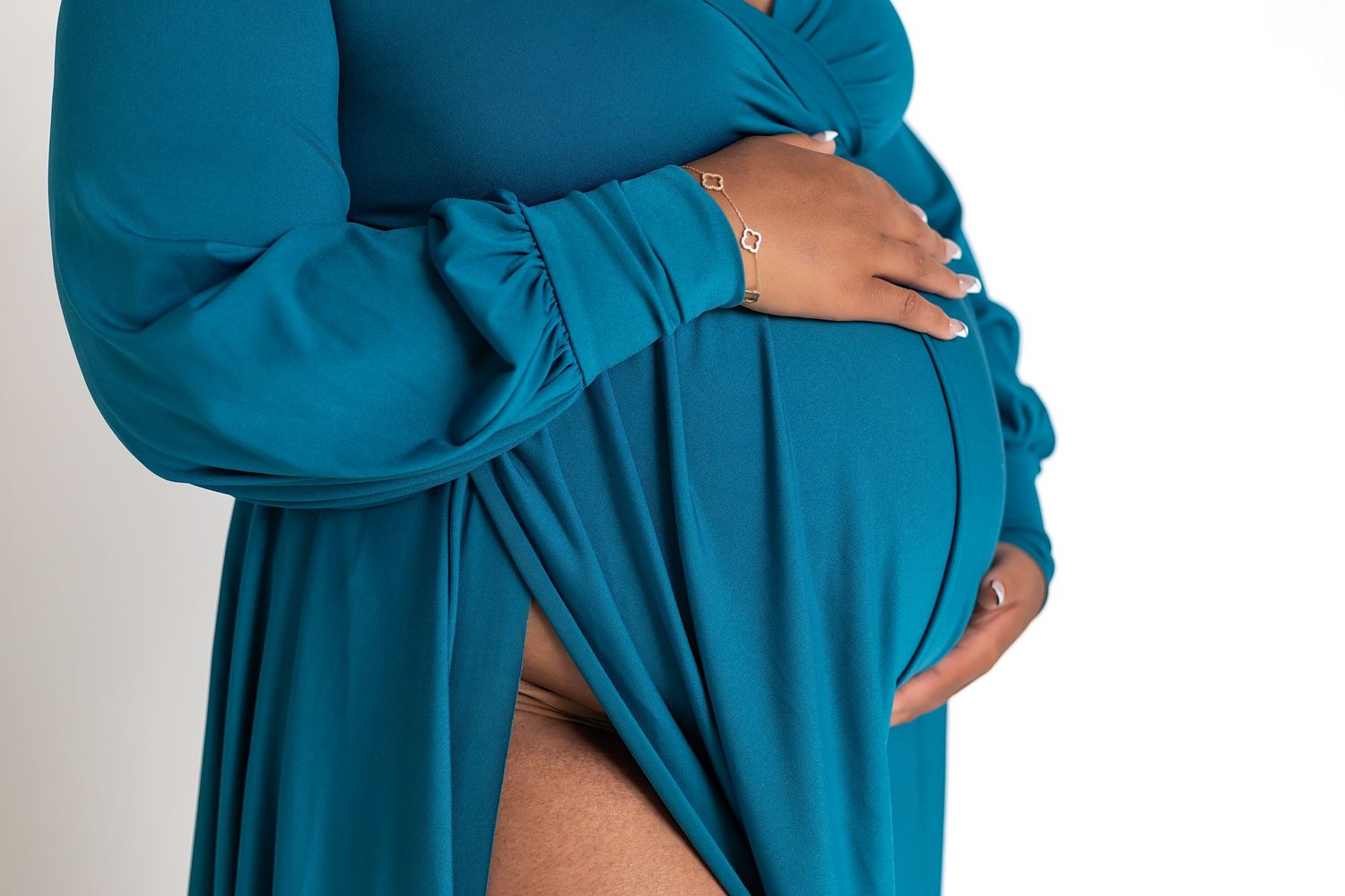 A pregnant woman in a teal blue dress gently holds her belly against a white background.
