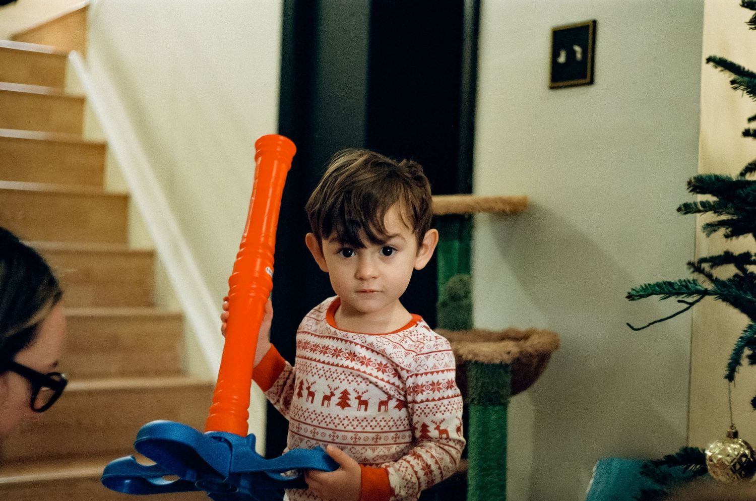 Child holding orange toy near Christmas tree and staircase.