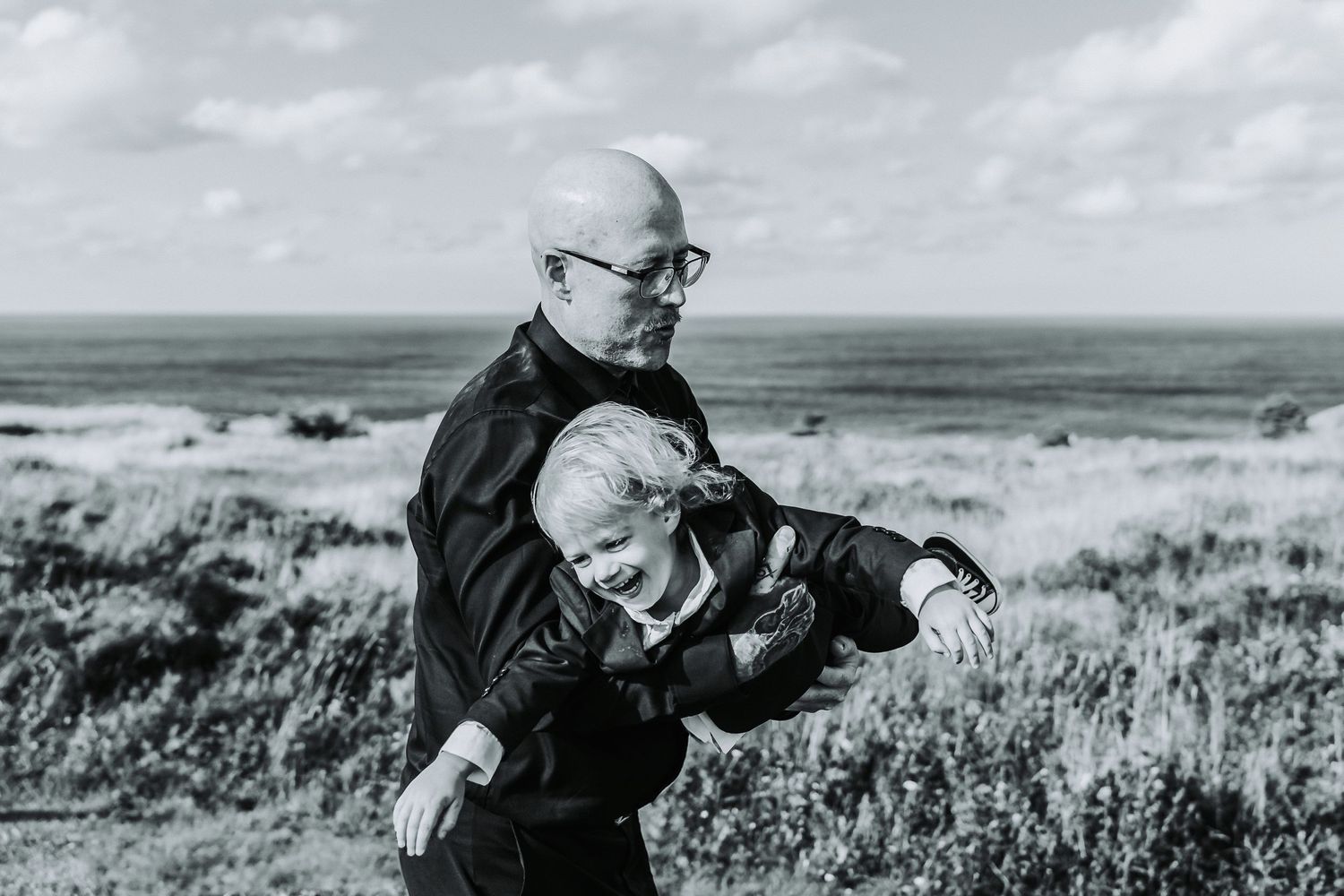 A black and white photograph shows a person in dark clothing swinging a child around like an airplane while standing in an open field.