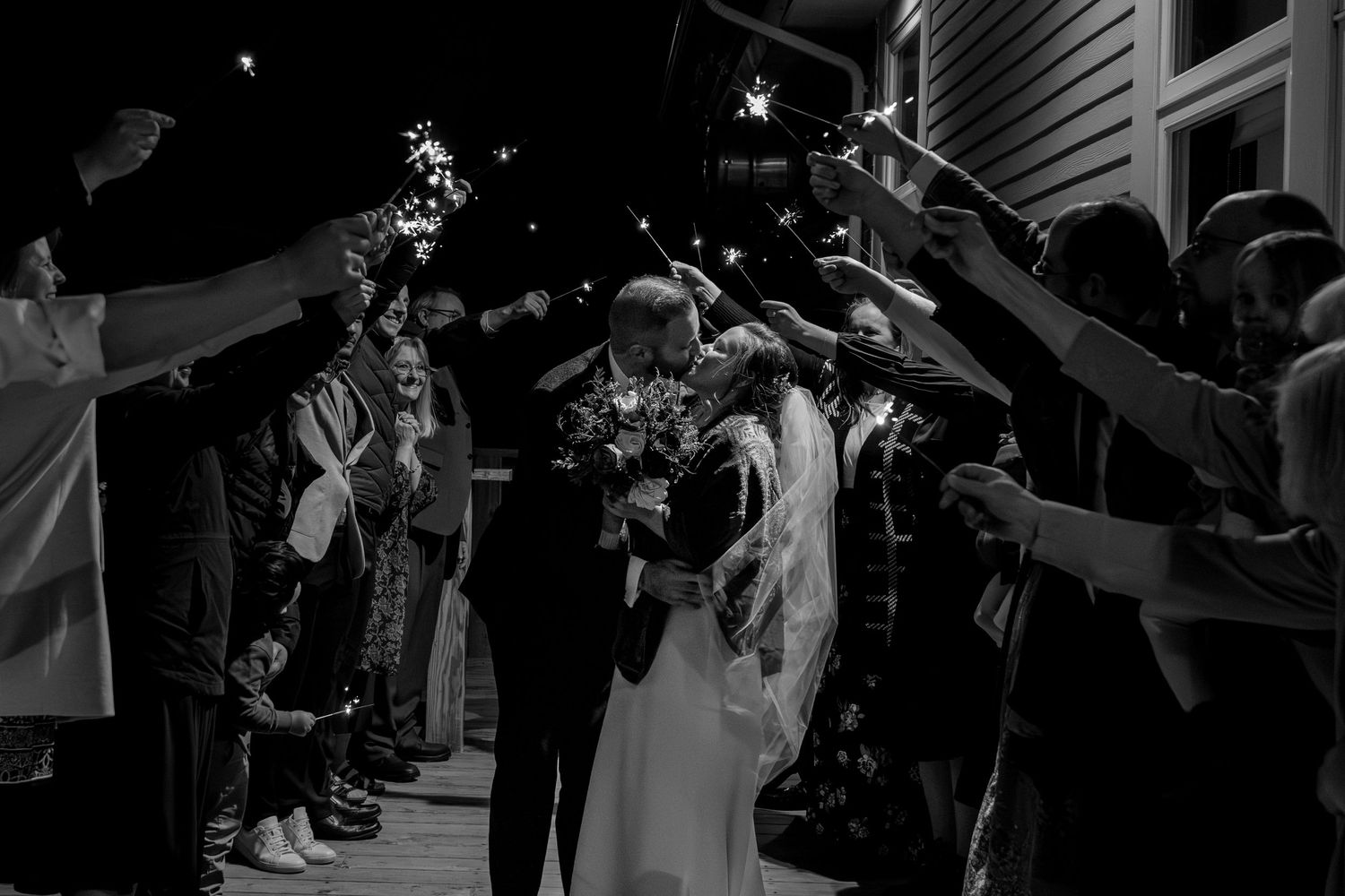 A gathering of wedding guests hold sparklers forming an illuminated archway as a couple exits in a black and white photo.