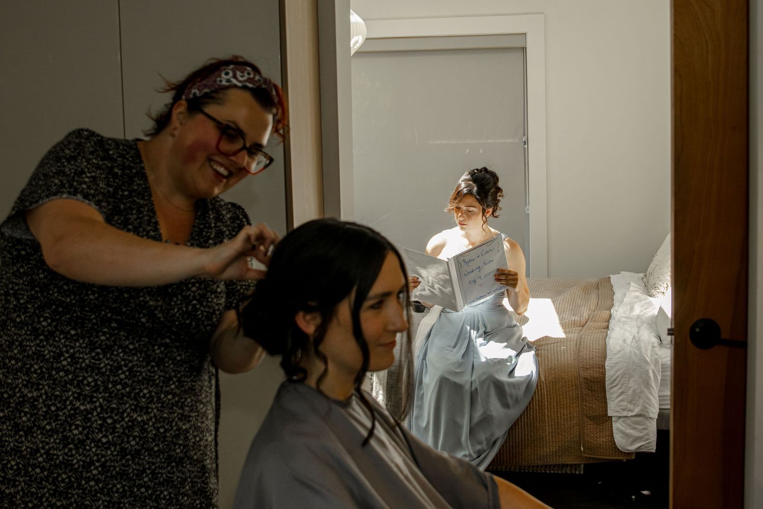 Behind-the-scenes photo of wedding preparations showing a hairstylist styling someone's hair while another person looks at a book