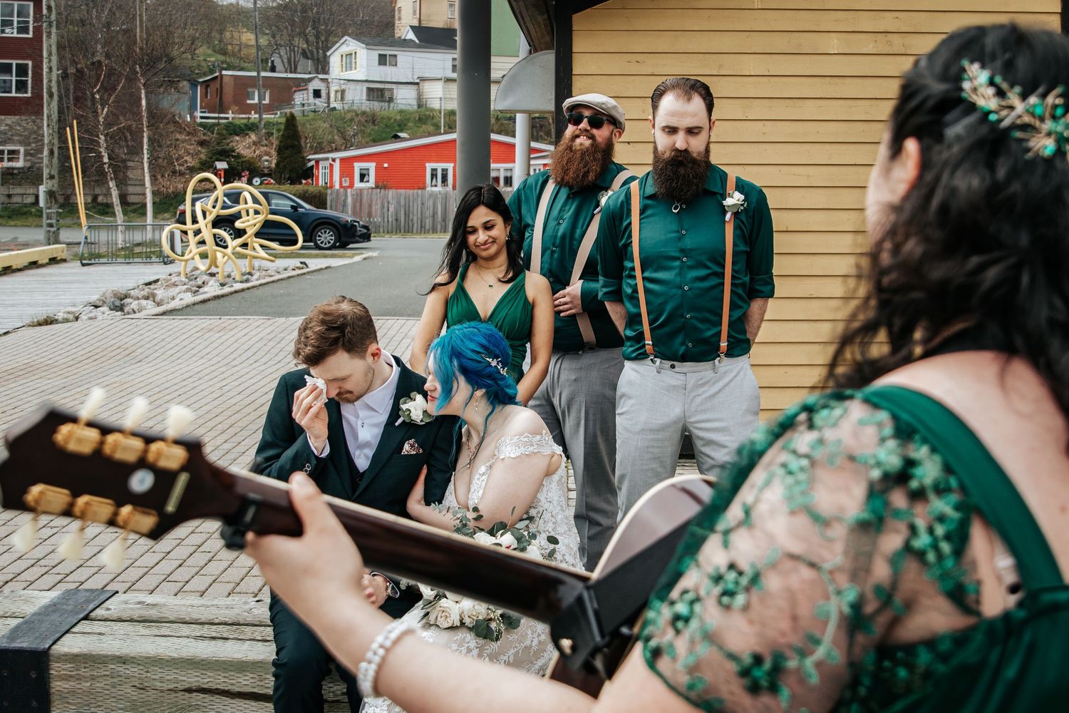 Wedding guests gather around a guitar player outdoors near wooden buildings on a casual autumn day, groom sheds a tear.