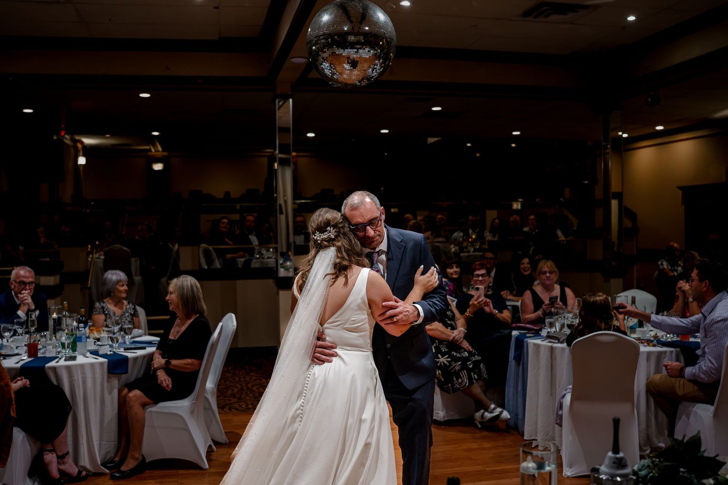 A father and daughter share a dance at a wedding reception with a disco ball overhead and guests watching from their tables.