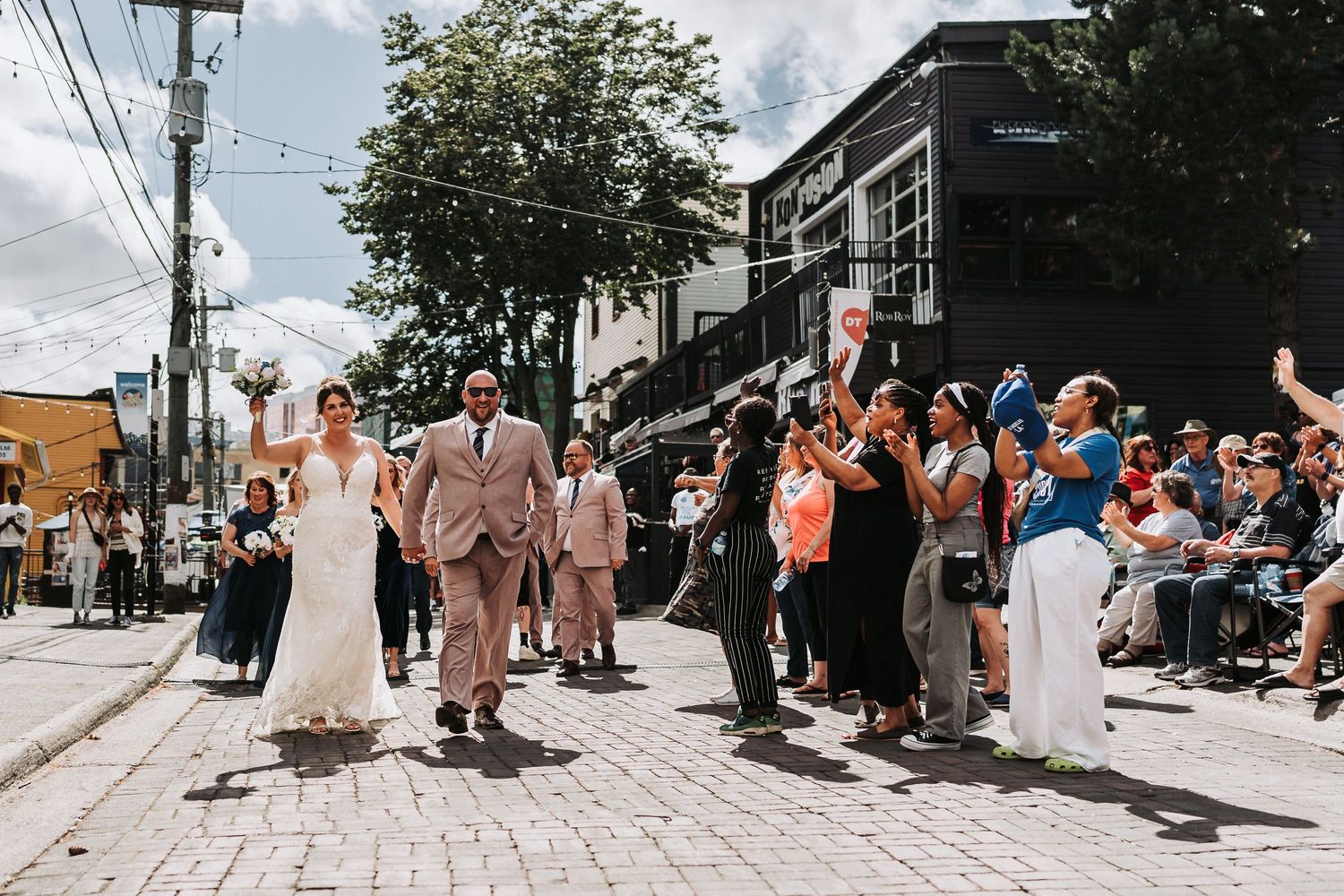 A wedding party celebrates on a lively urban street lined with buildings and spectators during a summer day.