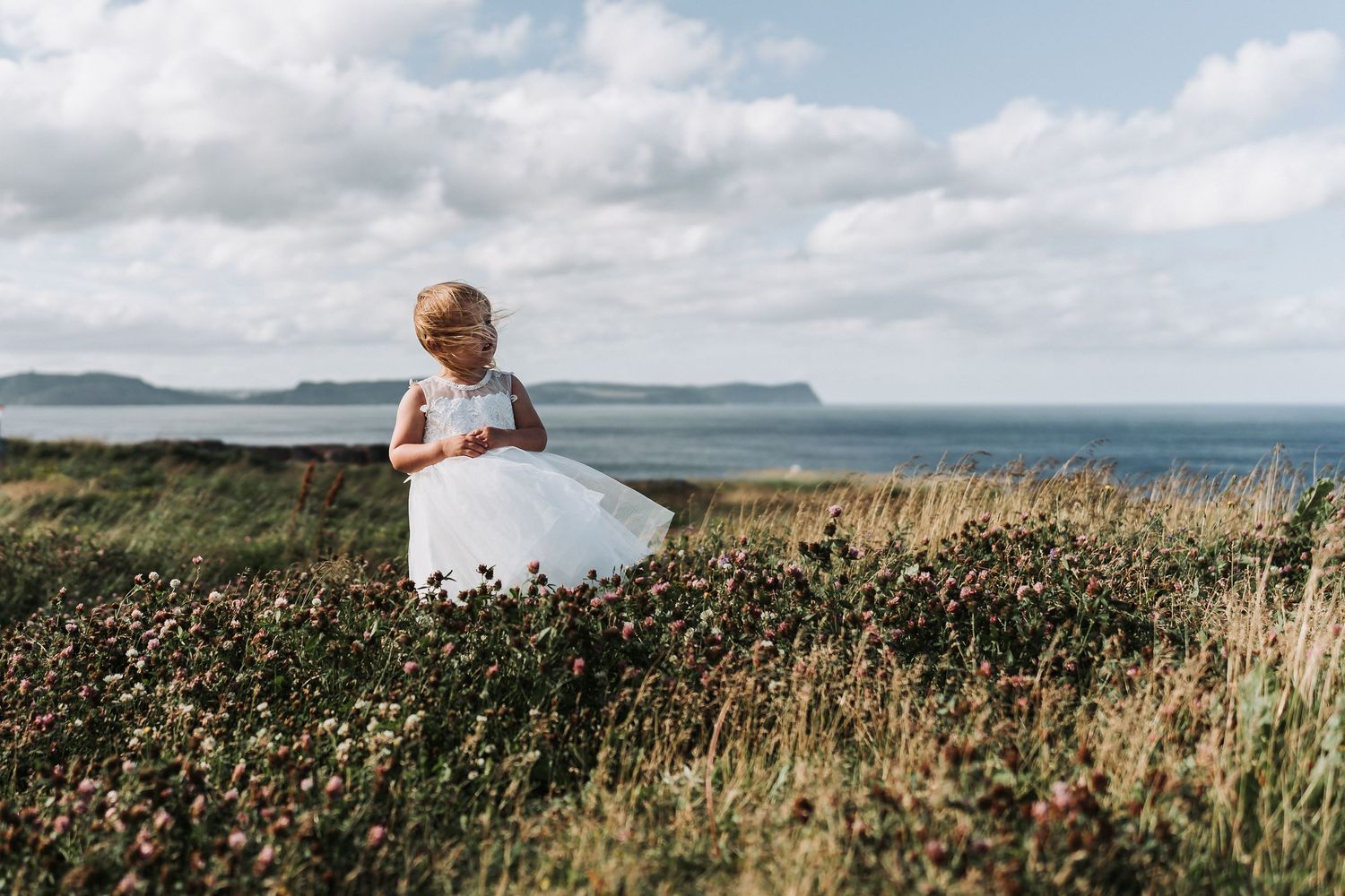 A small figure in a white dress stands amid golden grasses on a windswept coastal cliff overlooking distant ocean views.