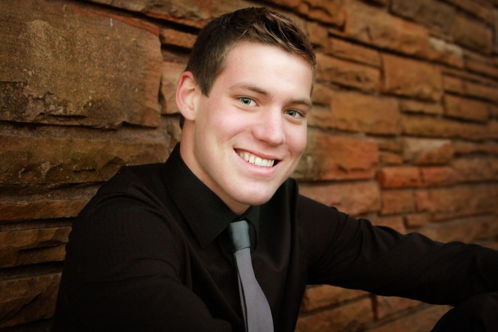 Young man in a black shirt and gray tie smiles against a rustic brick wall background