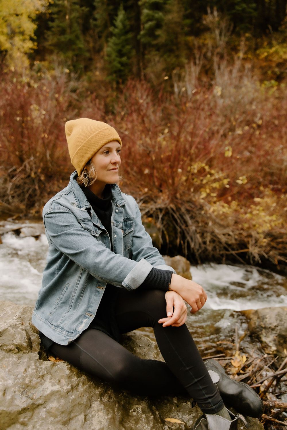 Casual autumn portrait of person in yellow beanie and denim jacket sitting on rocks near water.