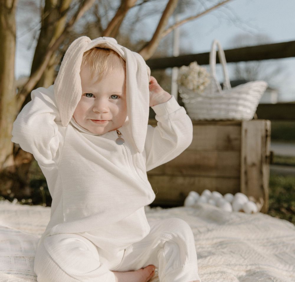 Playful baby wearing white bunny ears costume sitting on blanket outdoors.