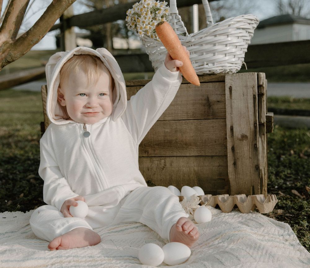 Sweet Easter scene with baby in white bunny costume sitting on blanket with eggs.