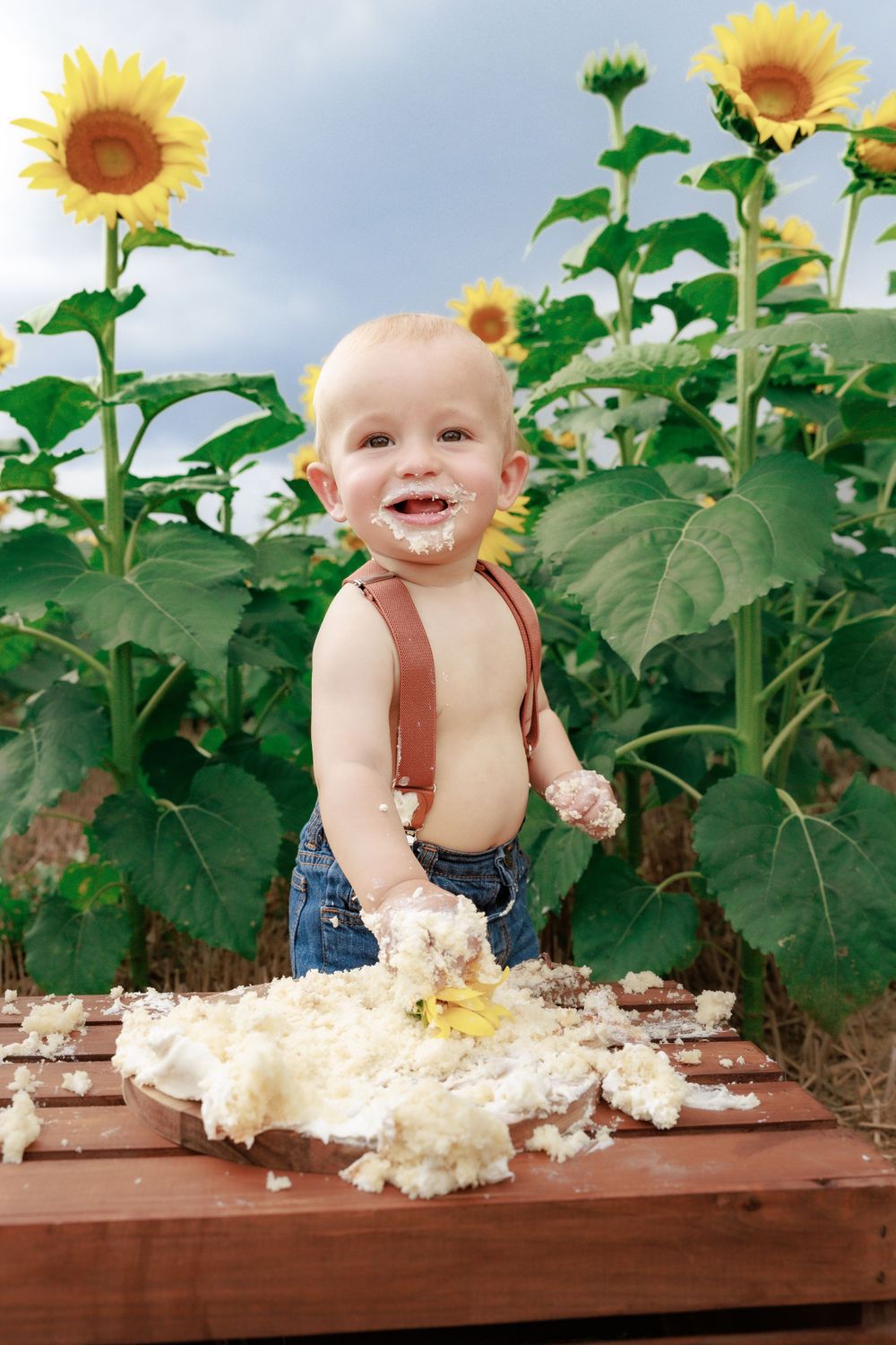 Happy toddler in overalls playing with cake in front of towering sunflowers.