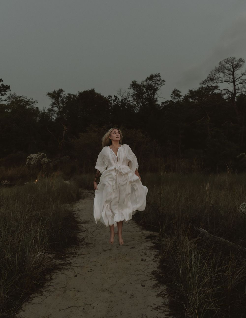 Ethereal scene of figure in flowing white dress running along sandy path at dusk.
