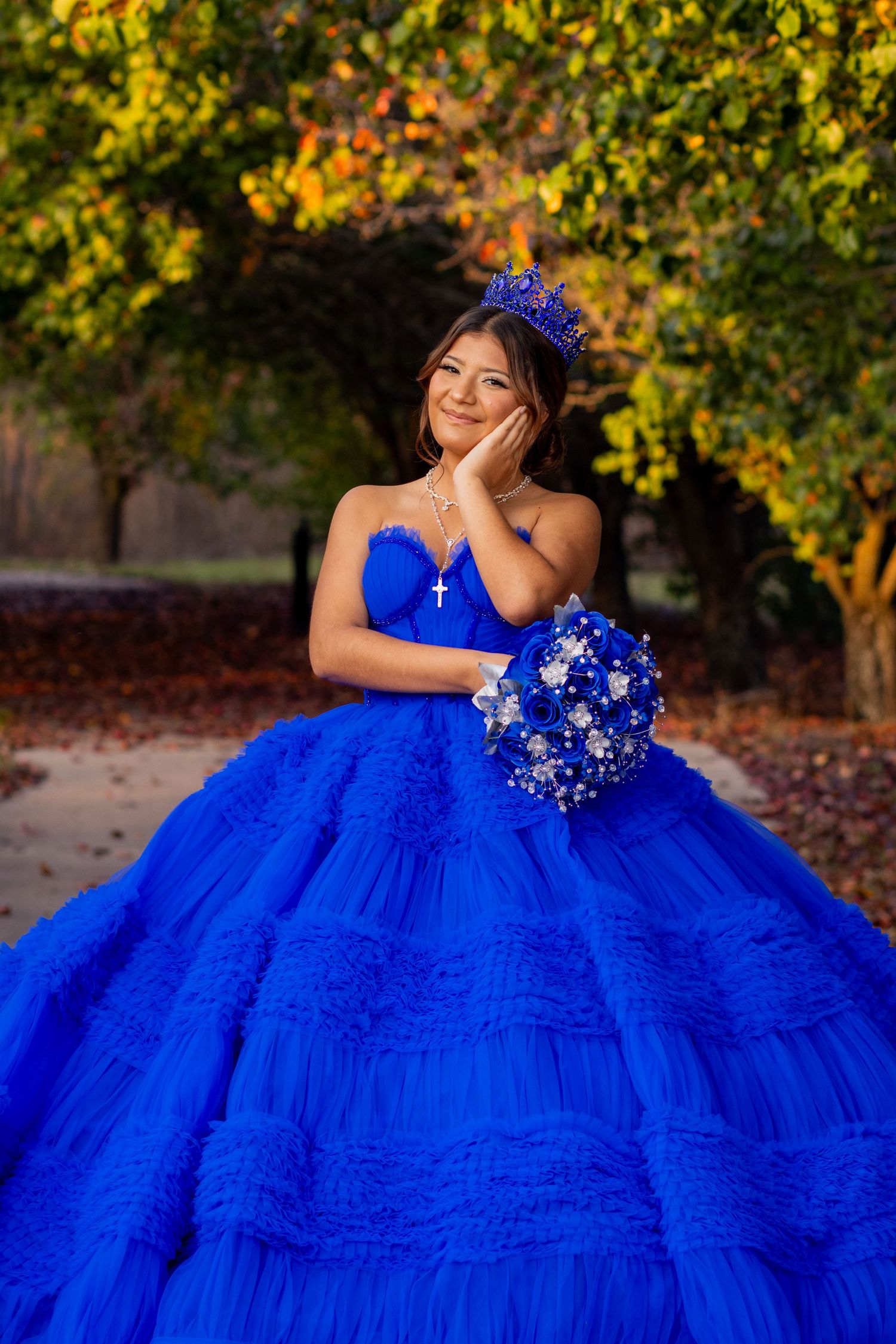 A vibrant royal blue quinceañera dress with ruffled layers photographed outdoors against autumn foliage.