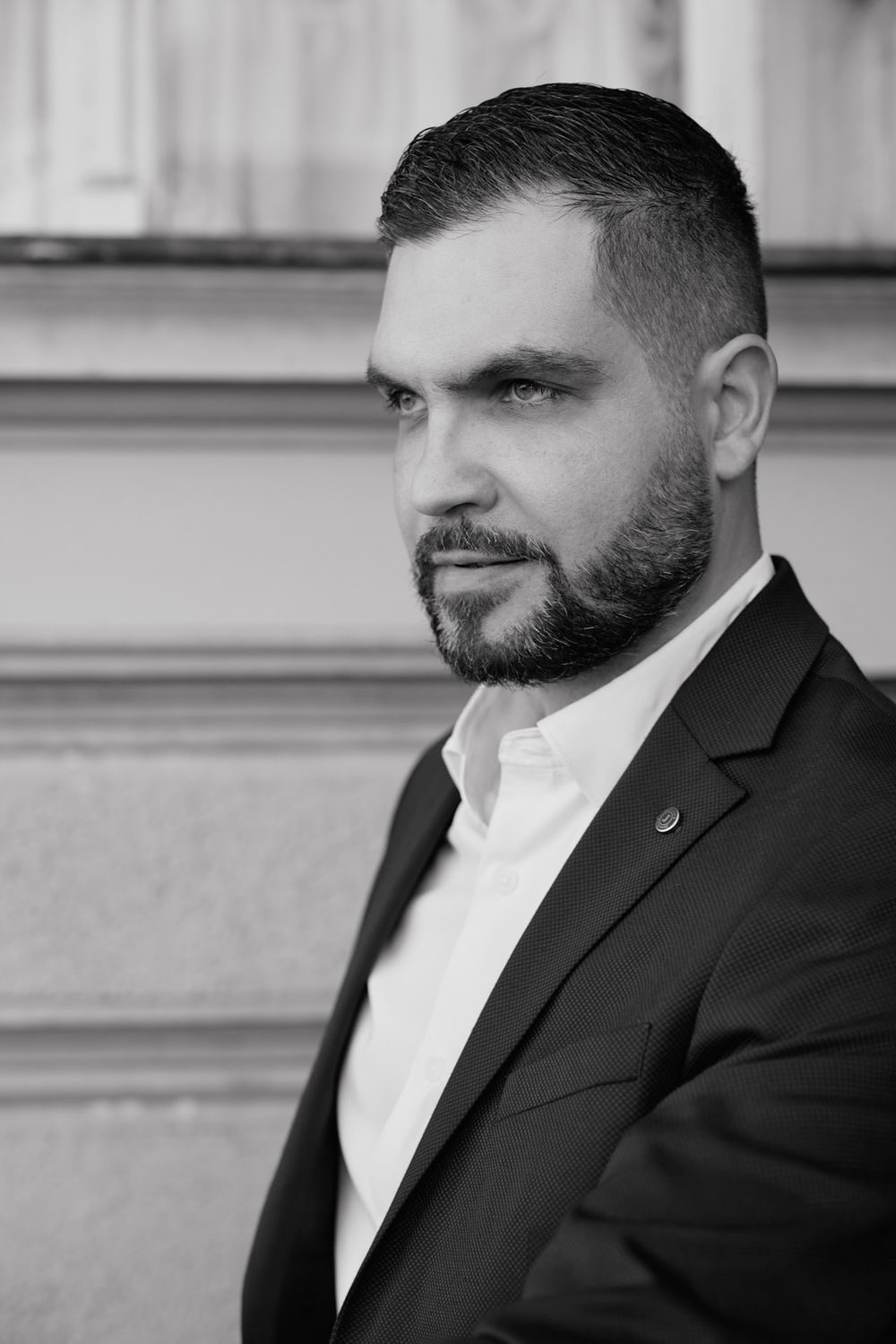Profile view of a person in formal attire with a dark beard against a light background.