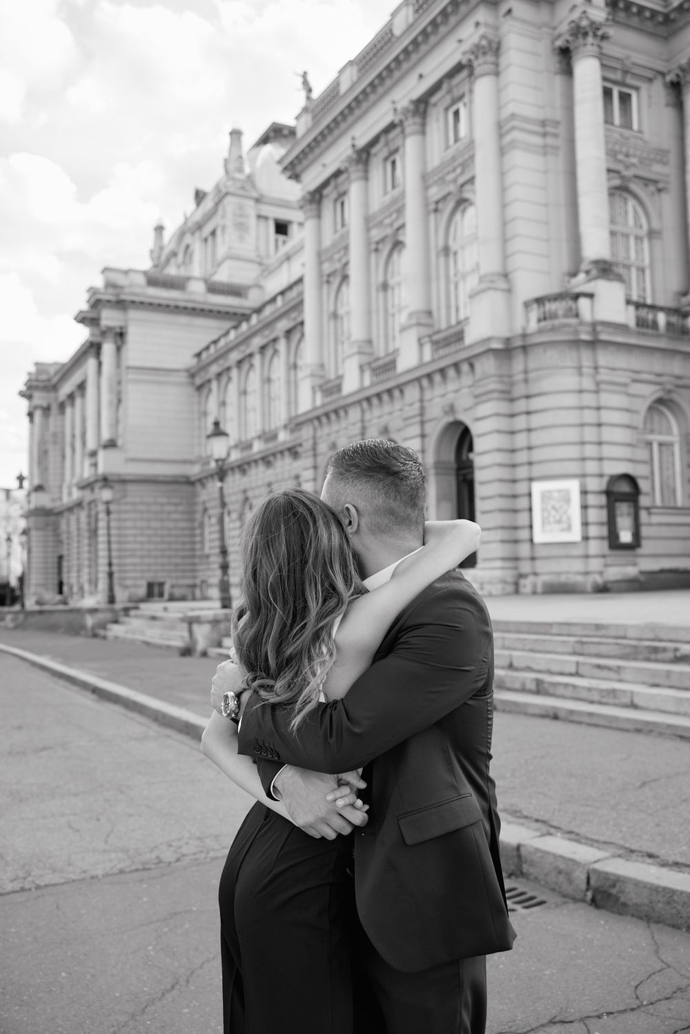 Couple embracing outside a classical European building with ornate stone facade and steps.