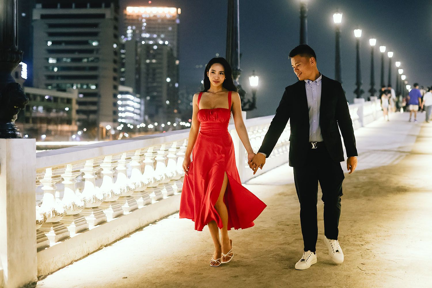 Couples walk along an illuminated waterfront promenade at night with city lights glowing in the background.