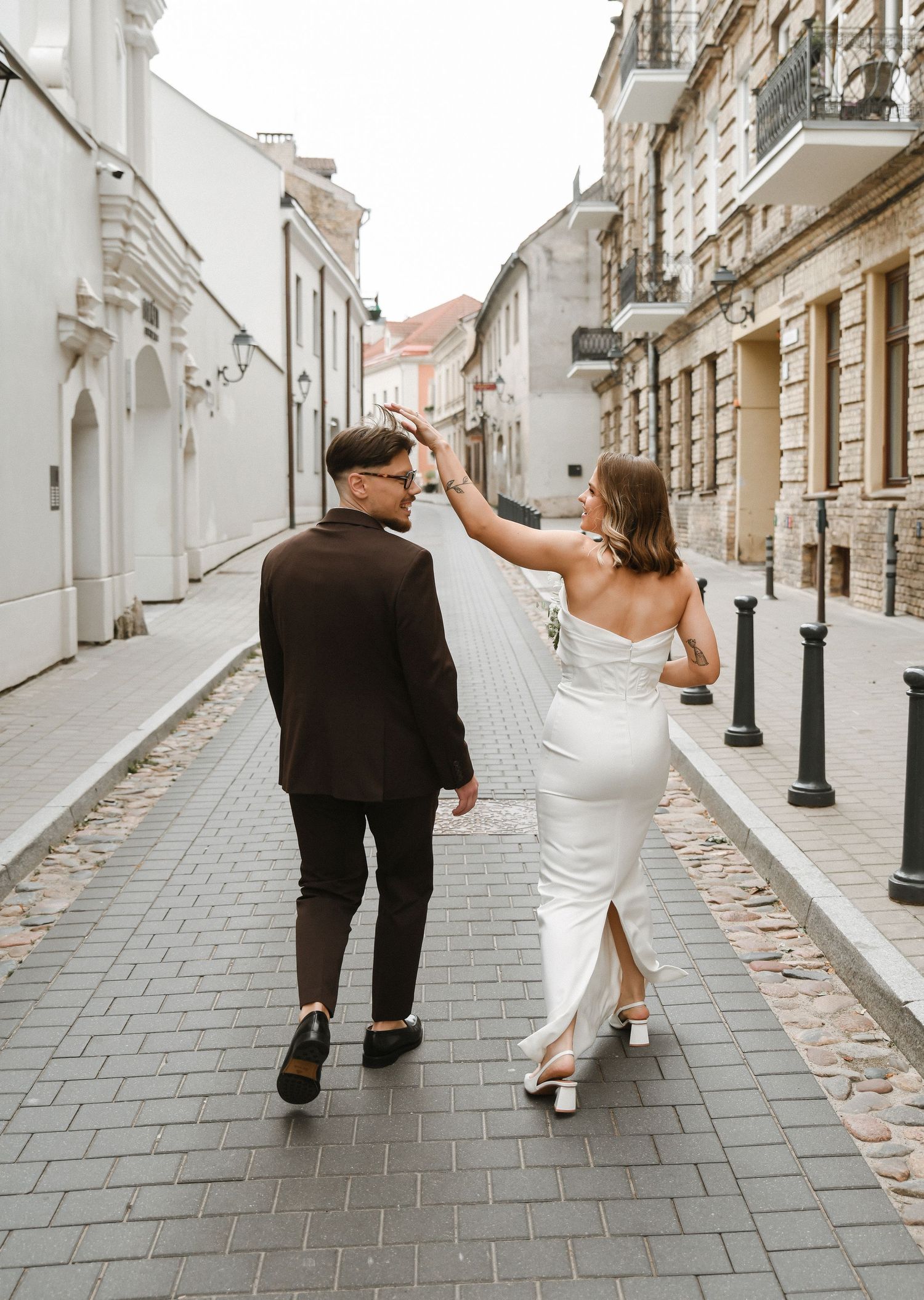 A couple walks down a charming European cobblestone street, with the bride in a fitted white dress and groom in black.