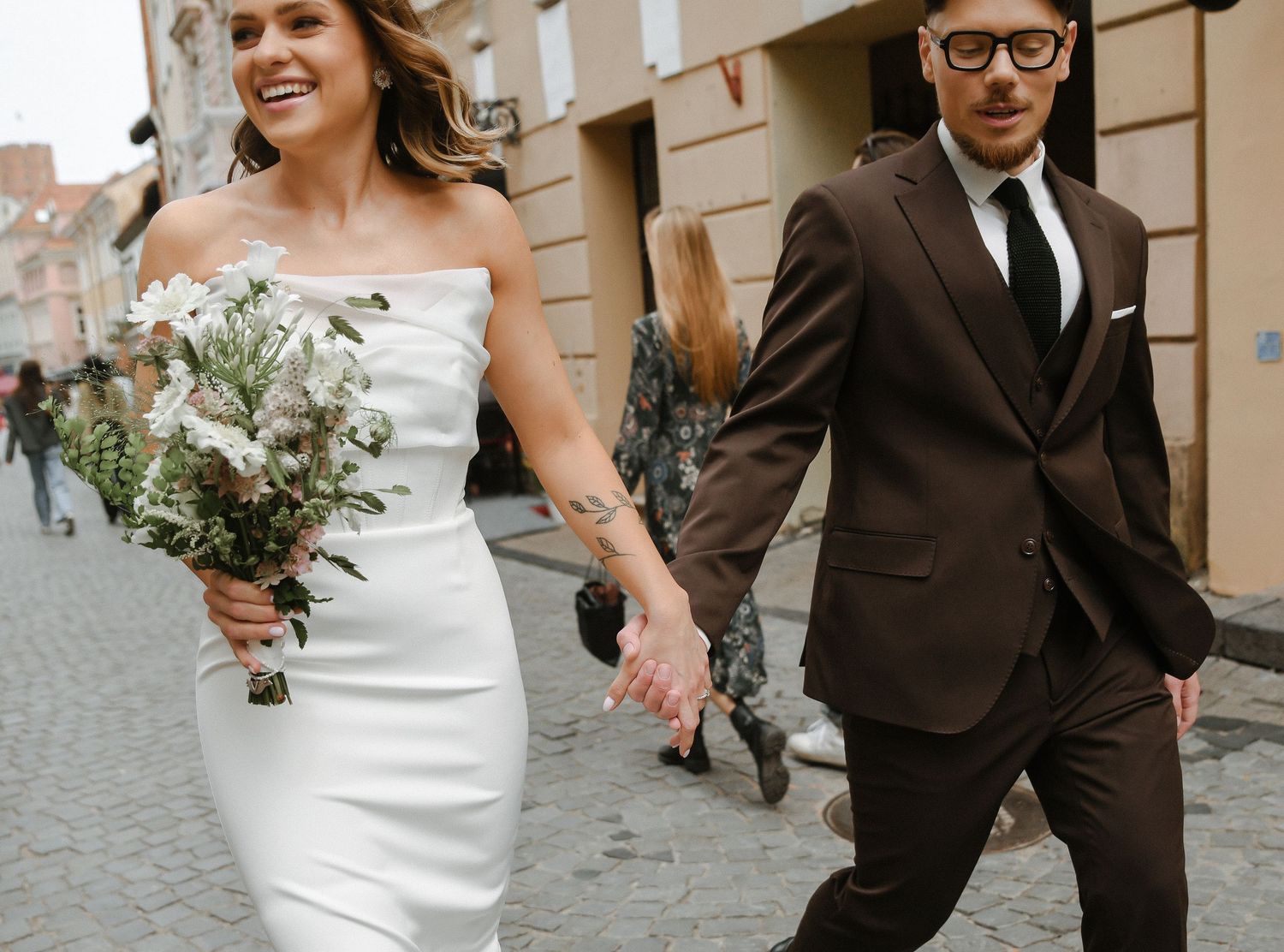 A wedding couple walks hand in hand along a cobblestone street while holding a rustic floral bouquet.