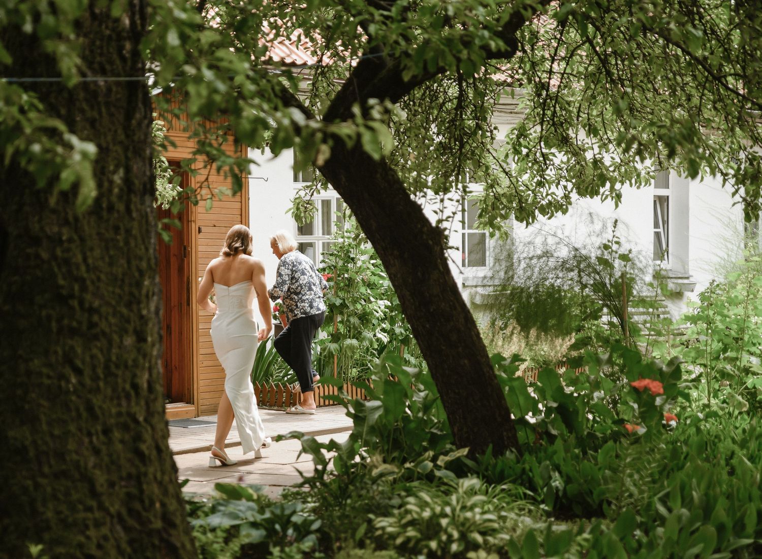 A figure in a white dress walks along a garden path beneath overhanging trees near a white building entrance.