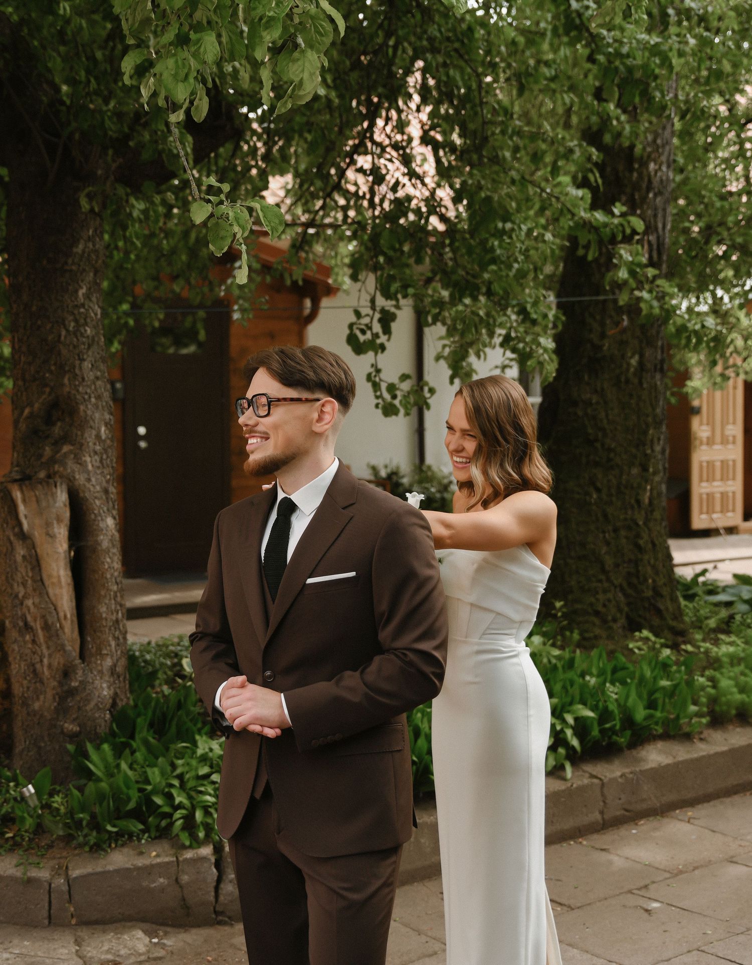 A couple in wedding attire shares a playful moment together on a tree-lined path.
