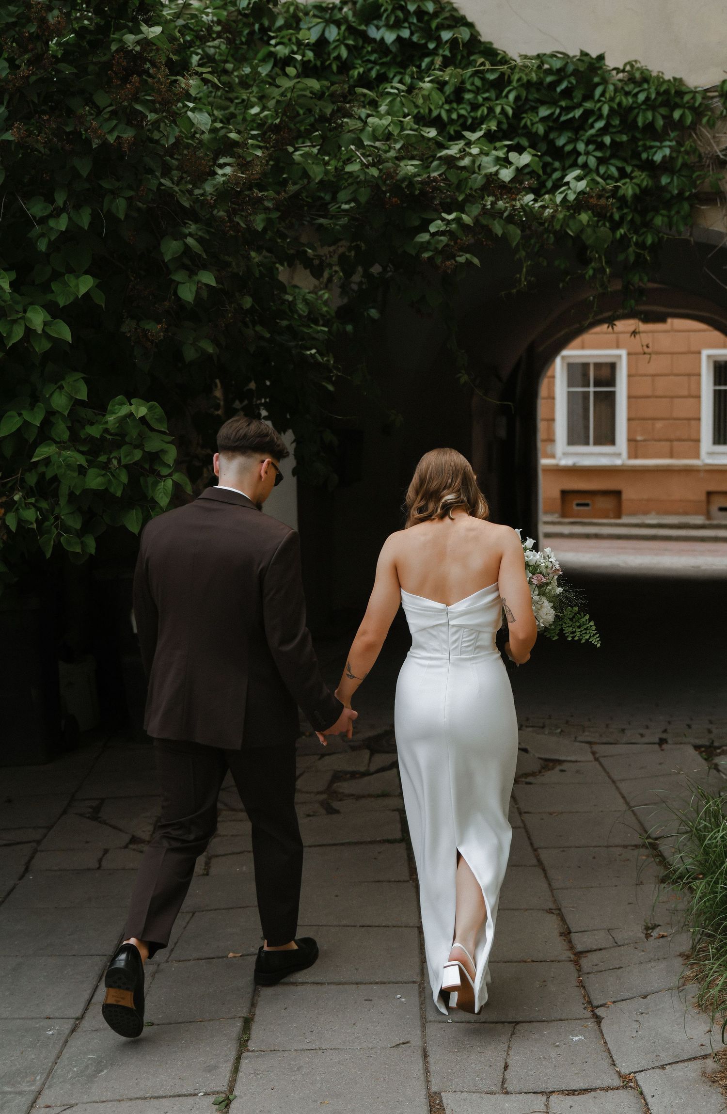 A couple in wedding attire walks together on a stone path through a garden setting.