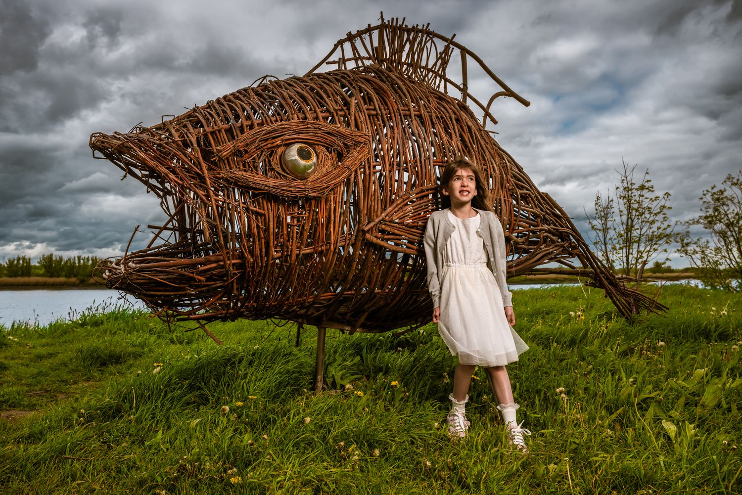 Meisje in witte jurk poseert bij groot houten kunstwerk in de natuur – creatieve communie fotografie door Bart Boodts