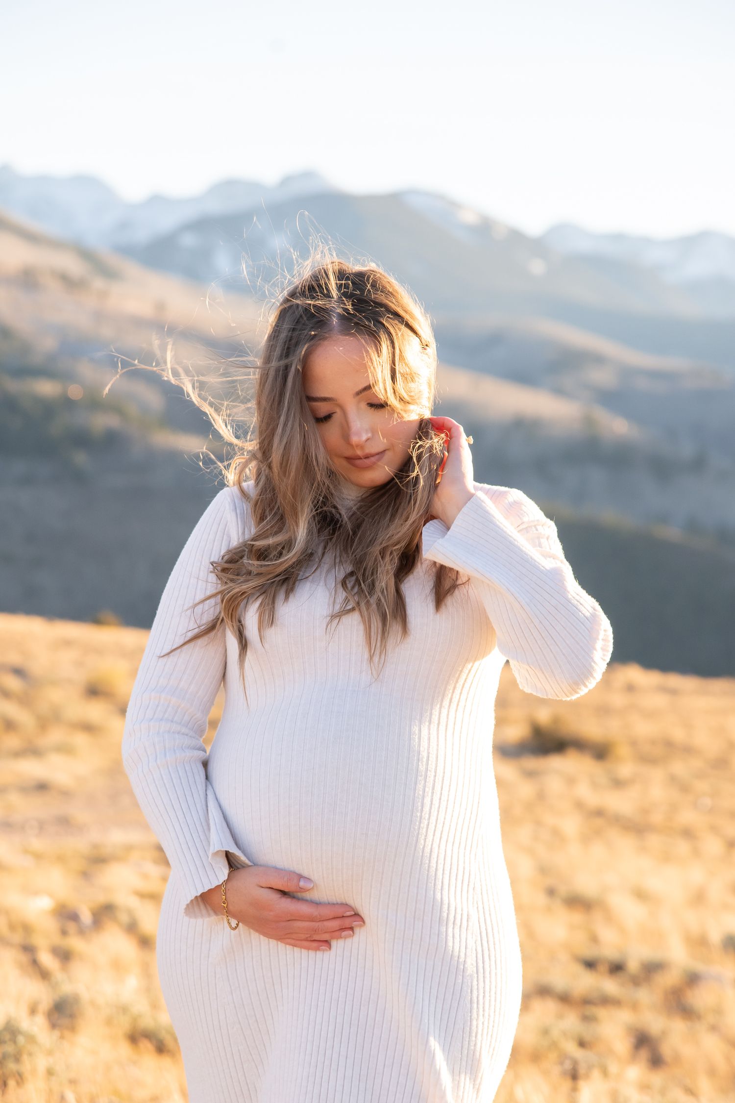 A fashionable woman in a cream sweater dress gazes at her pregnant belly with mountains in the background during Vail maternity photos