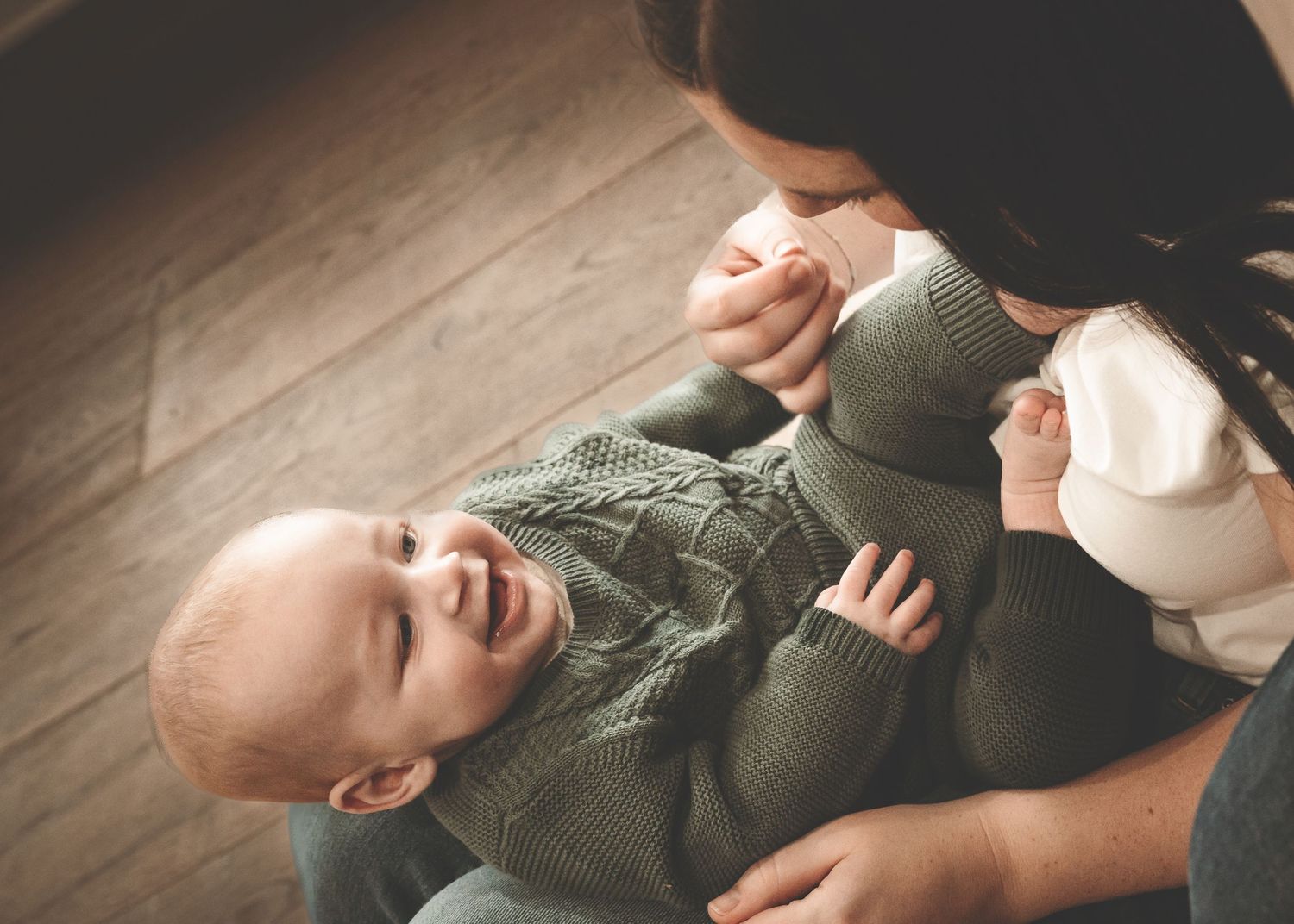 A baby in a green sweater lies on a wooden floor while being photographed from different angles during a lifestyle session