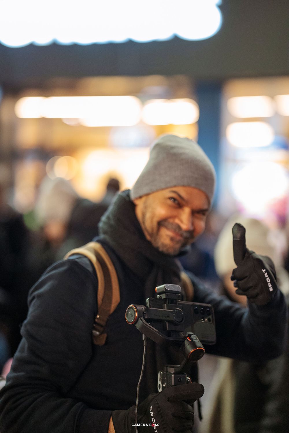 Photographer in winter clothing gives thumbs up while working with camera equipment in dark setting.