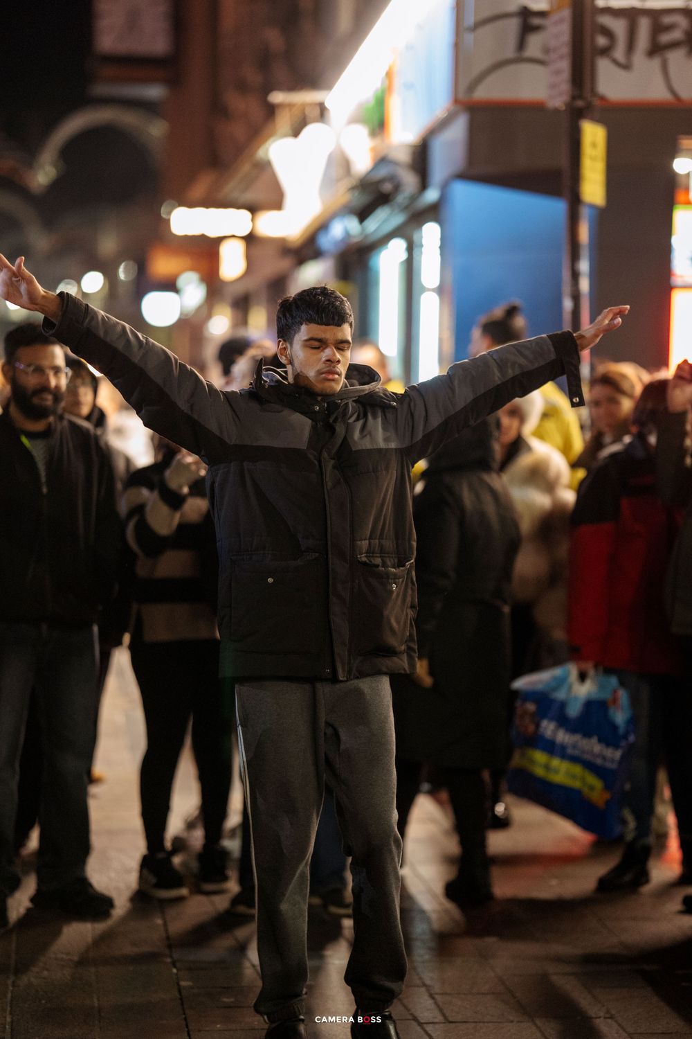 People with raised arms perform synchronized movements on a city street at night.
