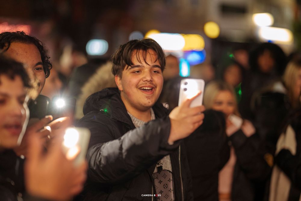 People in a crowd at night holding up phones with flashlights illuminated during an outdoor event.