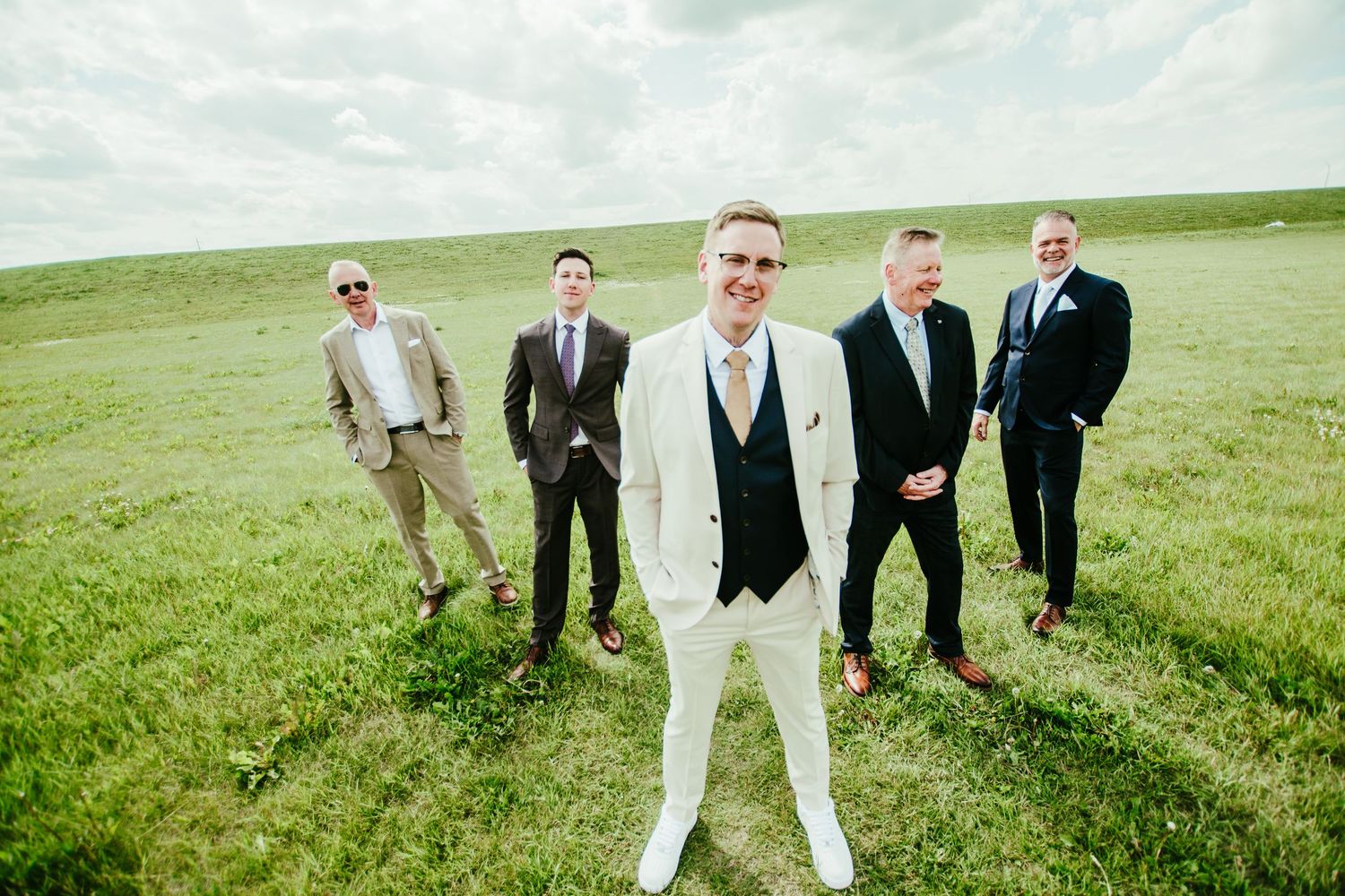 A group of formally dressed people walk across a green hillside meadow in a candid wedding party photo sequence.