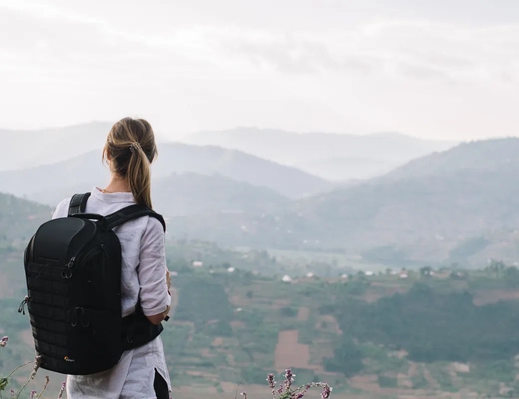 Person wearing a backpack stands on a hilltop overlooking a misty mountain landscape with distant buildings.