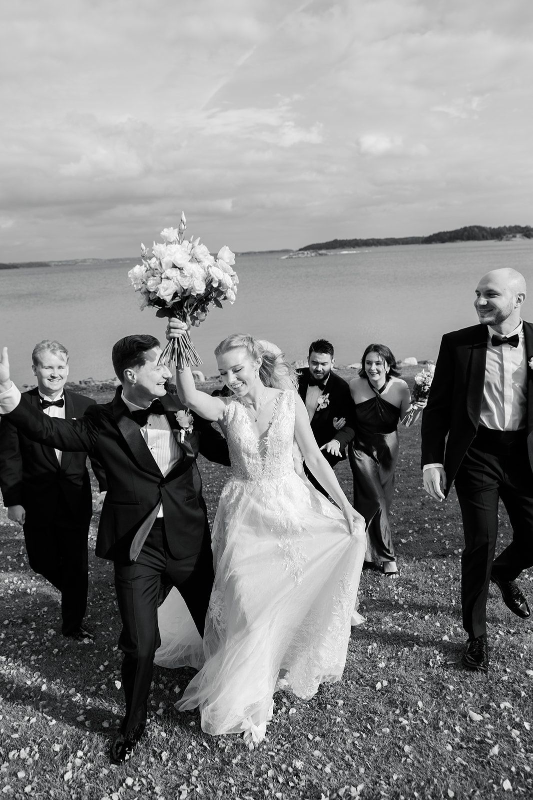 Wedding party dances joyfully together in a line across a scenic coastal meadow in black and white.