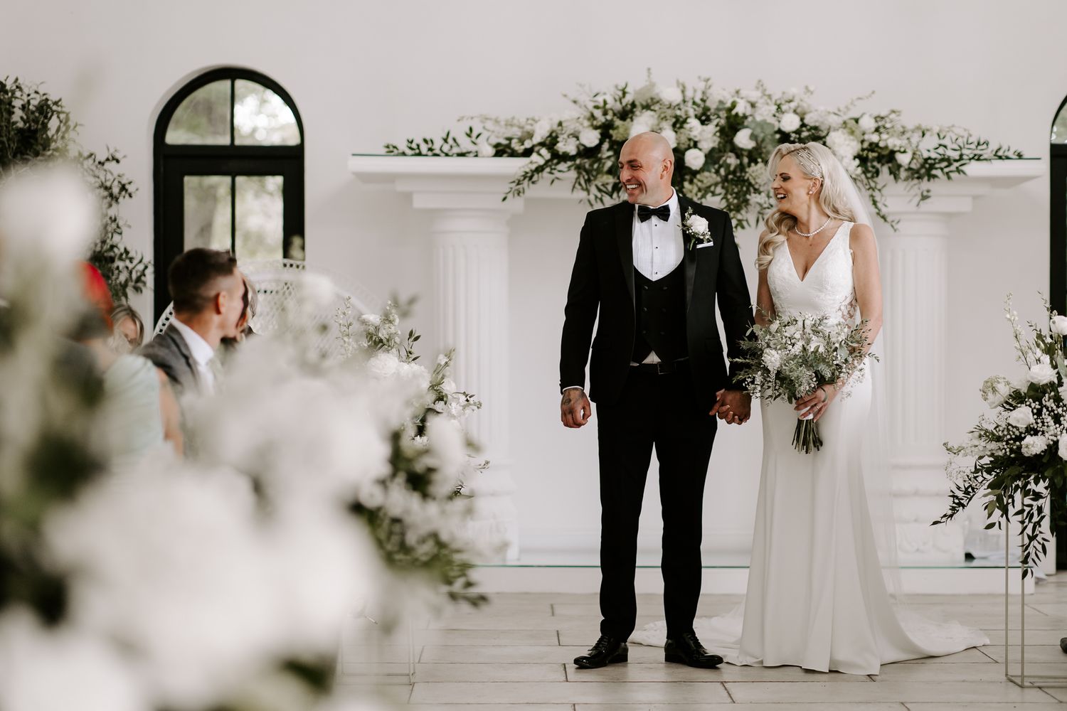 Elegant wedding party poses in a bright white room adorned with delicate floral arrangements and greenery.