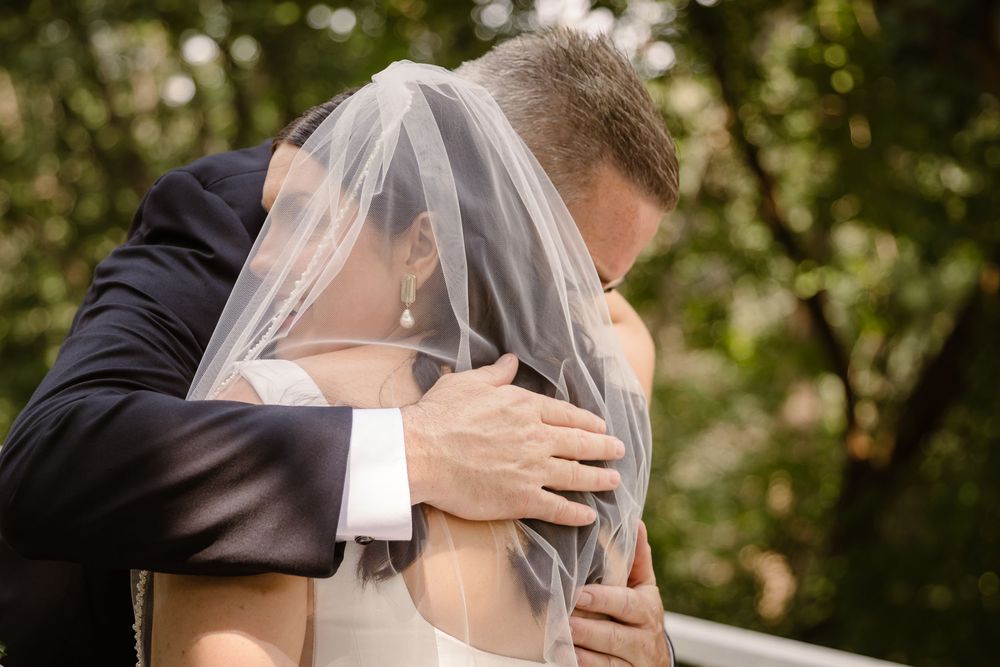 A romantic wedding couple embraces and kisses outdoors surrounded by soft bokeh lights and greenery.