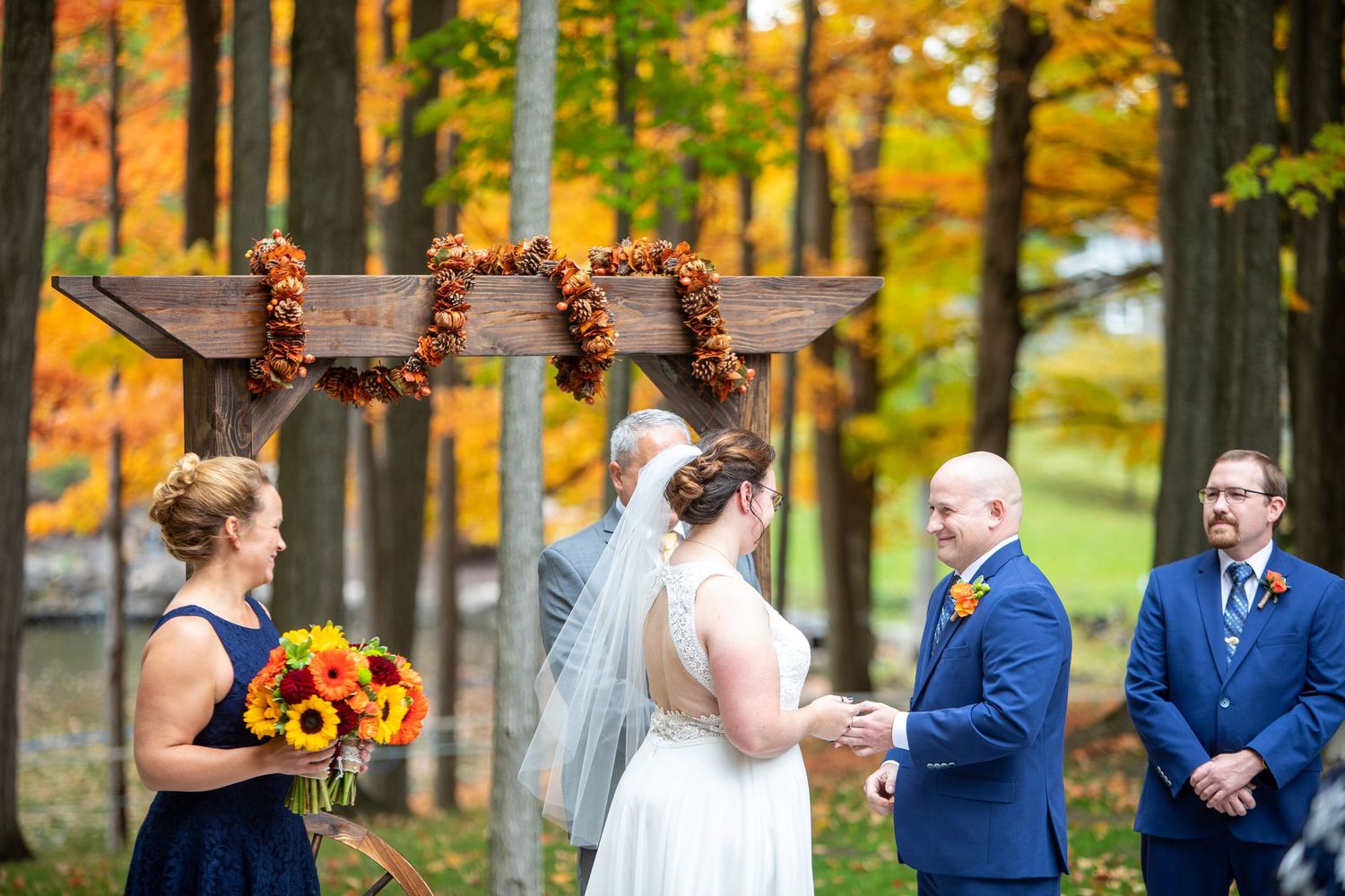 Outdoor autumn wedding ceremony under wooden arch decorated with fall foliage and sunflowers in forest setting.