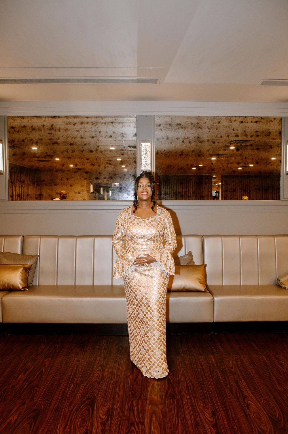 A person in a sparkly gold sequined dress poses against a backdrop of white wainscoting and dark hardwood floors.