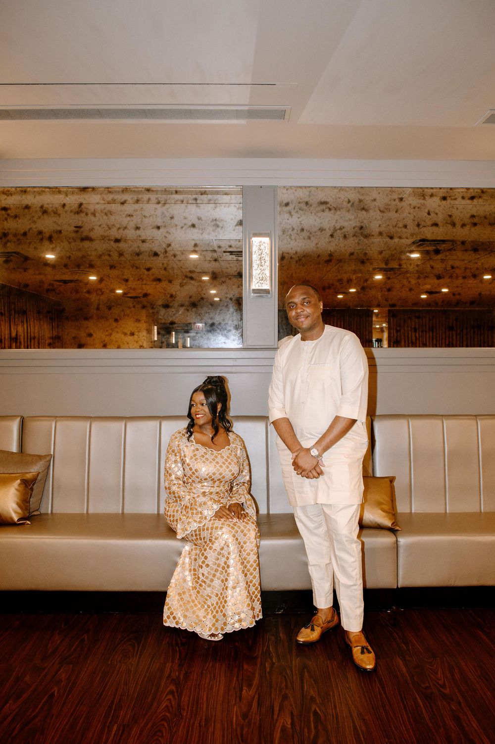 A couple poses together in a stylish indoor venue with white bench seating and wooden floors.