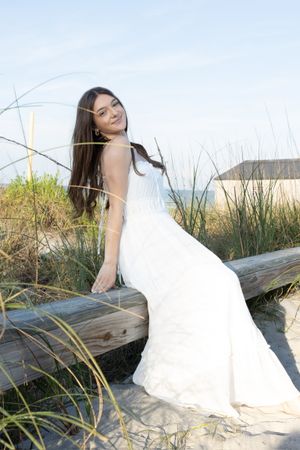 Woman in white dress posing on wooden bench surrounded by beach grass.