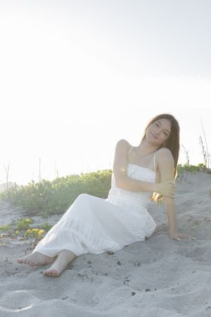 Woman in white dress resting on sandy beach during sunset.